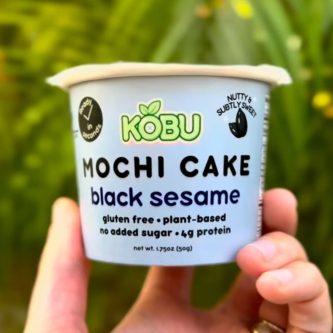 Hand holding a black sesame KOBU cake cup against a green plant background